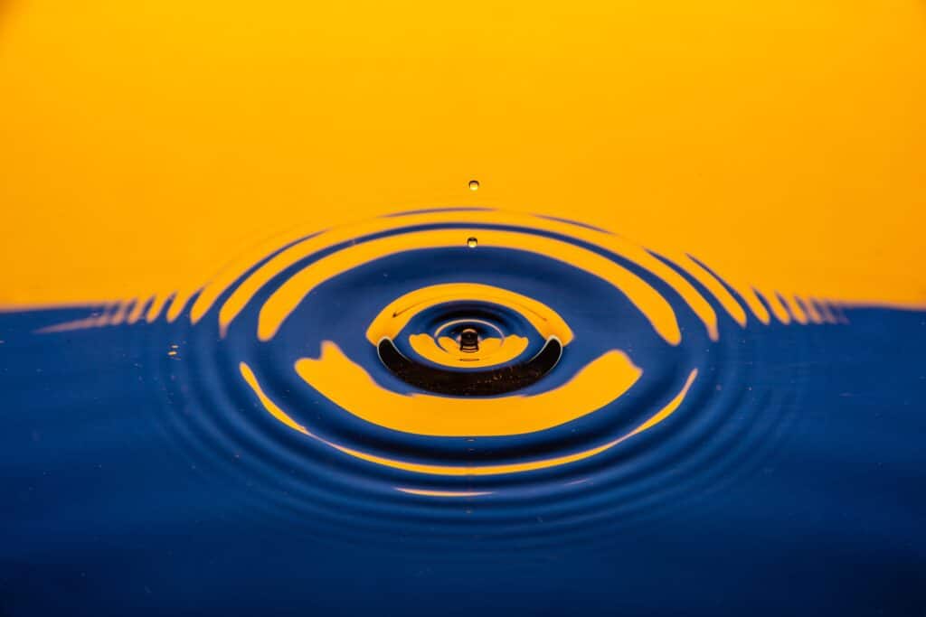 A drop of water hits a still, deep blue body of water with a contrasting yellow reflection towards the top of the image